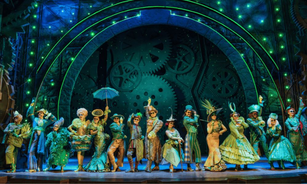 Best West End shows in London: This is an image of the Wicked cast dancing in the middle of the stage with green costumes and lighting, with cogs on the screen behind them.