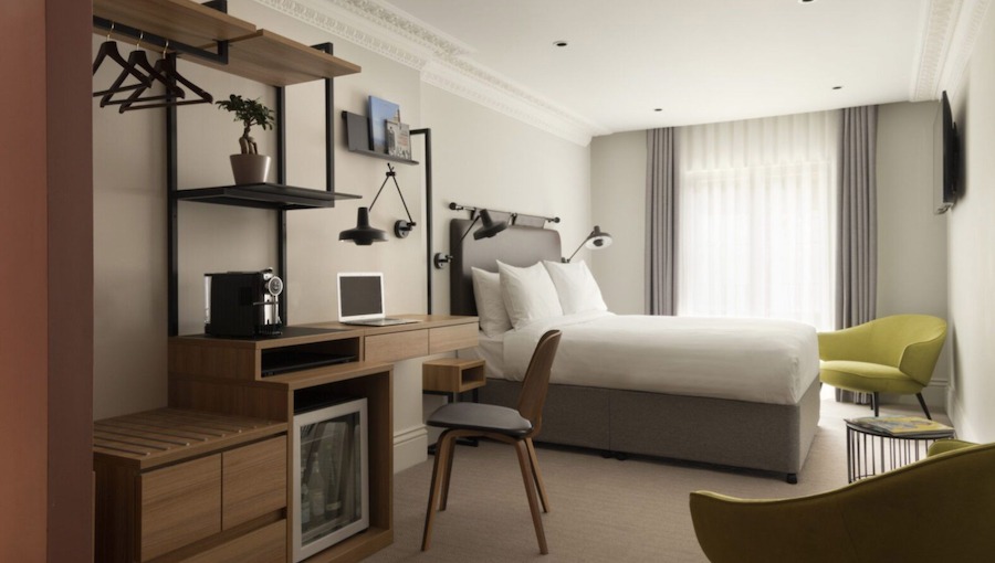 This is an image of a hotel bedroom with a double bed that is neatly made, wooden furnishings and natural daylight coming in from the windows by the bed.