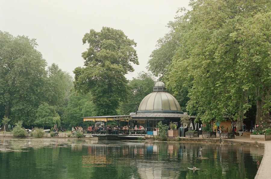 This is an image of a beautiful park with green trees, a cafe and a pond with still water.