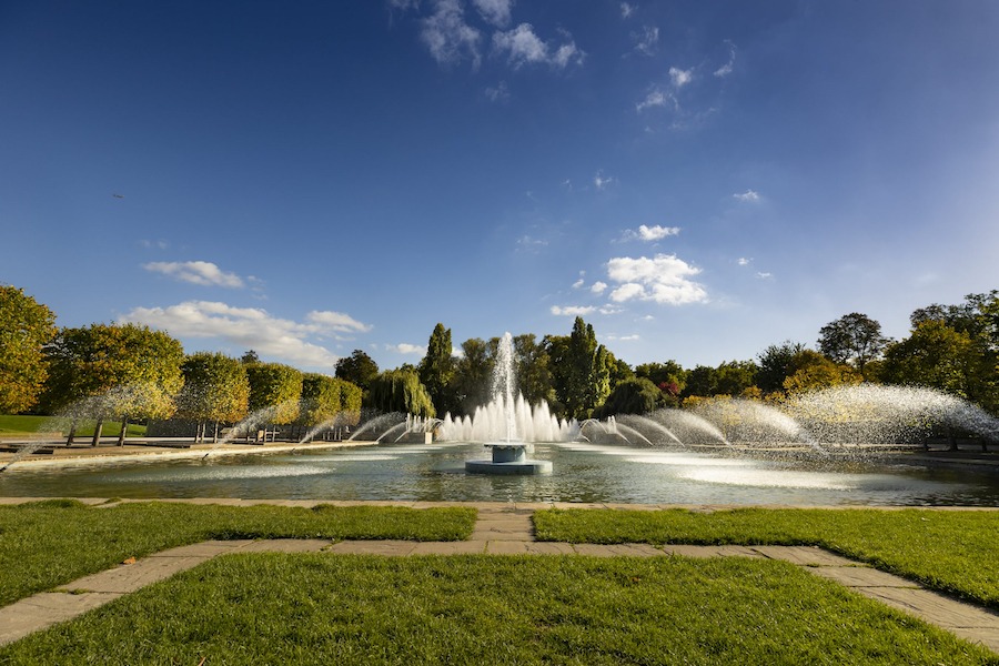 This is an image of an open space in a green park. There is a pretty water fountain in the middle of the park and blue skies above.