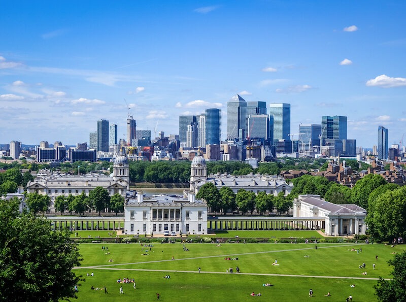 This is an image of a green park with an observatory in the background and bright blue skies. We can see the skyline of London in the far background.
