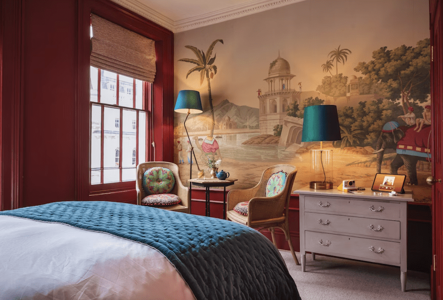 This is an image of a boutique hotel with a big double bed. There is an interesting painting on the wall opposite the bed of a nature scene with a beach and camels. The room has subtle hints of red in the furnishings that makes it vibrant and pleasing.
