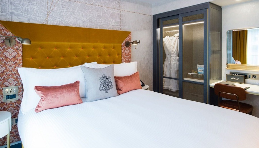 This is an image of a stylish hotel with a big double bed and white bedding, soft pink cushions and a plush velvet dark gold headboard behind the bed. There is also a glass wardrobe with lighting inside where a bathrobe is hanging.