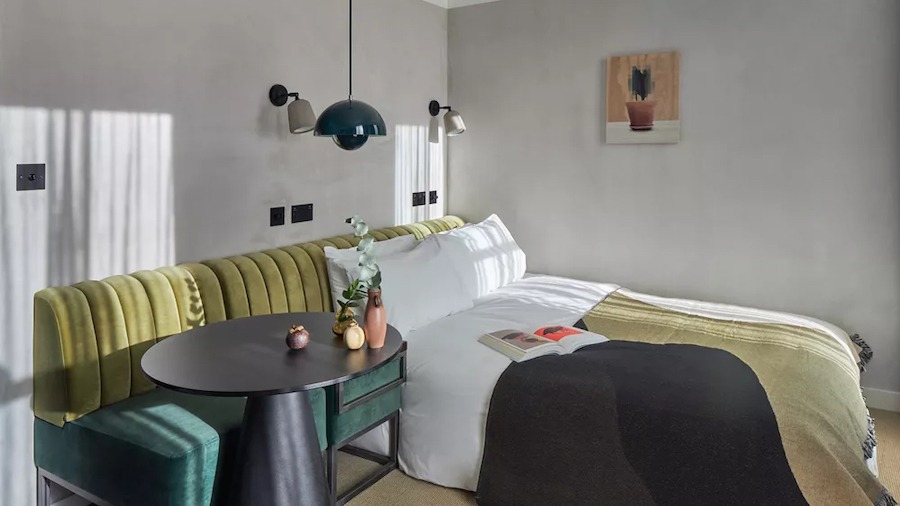 This is an image of a hotel bedroom with a double bed and minimalistic furniture. The room is toned in neutral colours and has subtle muted touches of green. There is also pleasing dappled sunlight shining in the room.