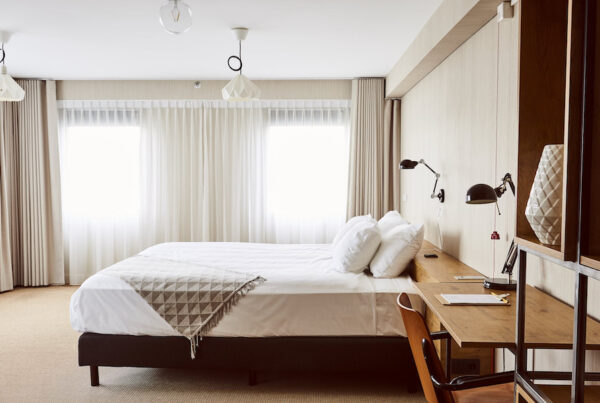 This is an image of a bright hotel room toned in muted colours, like beige, taupe, soft brown and white. There is a big double bed in the middle of the room and a wooden desk and chair beside the bed with a black table light.