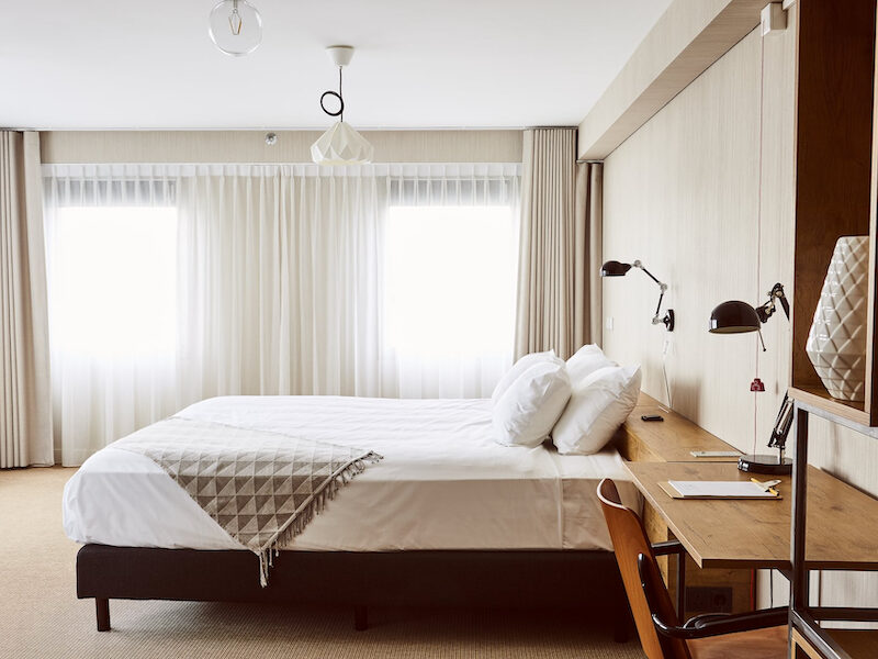 This is an image of a bright hotel room toned in muted colours, like beige, taupe, soft brown and white. There is a big double bed in the middle of the room and a wooden desk and chair beside the bed with a black table light.