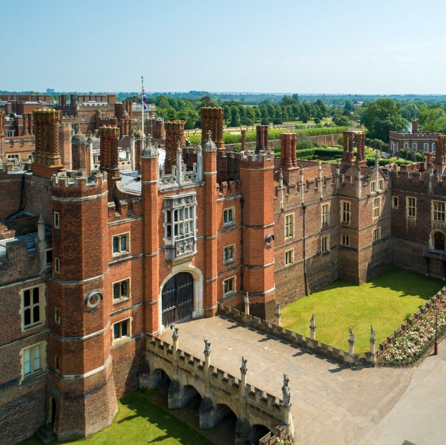This is an image of Hampton Court Palace. The gardens around the palace are impeccably kept and the sky is bright and blue.