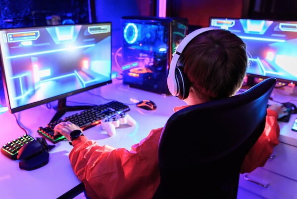 This is an image of a boy playing games on his computers. He has multiple screens set up and they are emitting bright neon lights.