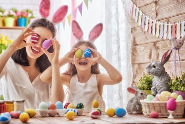 This is an image of a woman and a girl holding up easter eggs over their eyes. They are smiling and are wearing bunny ears headbands around their heads. There are lots of Easter Eggs on the table in front of them and the room is lit brightly.