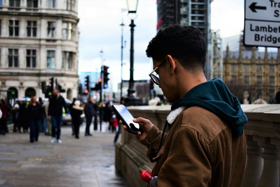 Young man wearing glasses and a brown corduroy jacket looking at his smartphone on a busy London street, with a Lambeth Bridge road sign and the Houses of Parliament visible in the background.