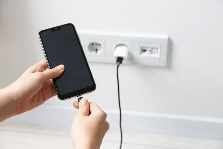 Person plugging a charging cable into a smartphone, with a wall adapter connected to a European-style power outlet in the background.