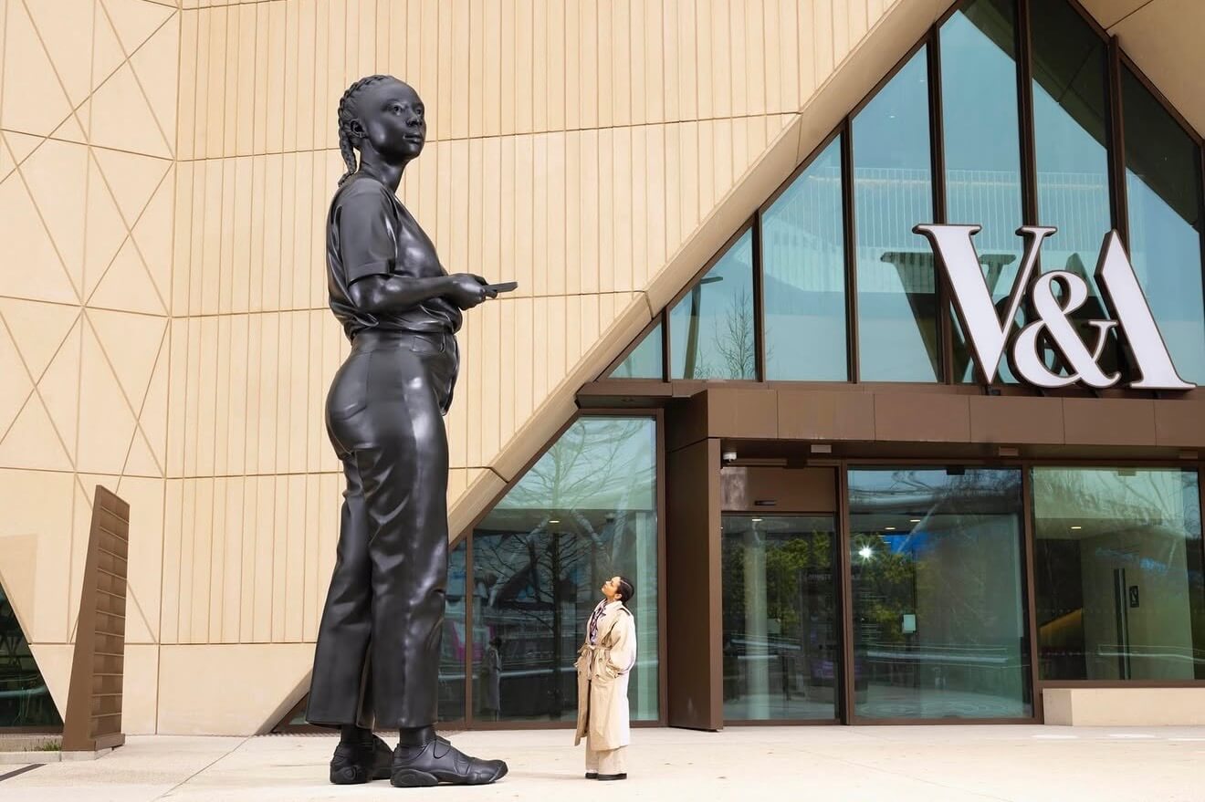 A towering dark bronze sculpture of a young Black woman in casual clothes and trainers stands outside the entrance of V&A East Museum in Stratford. A real person in a beige trench coat gazes up at the monumental figure, dramatically highlighting its scale against the museum's angular golden facade and large V&A signage.
