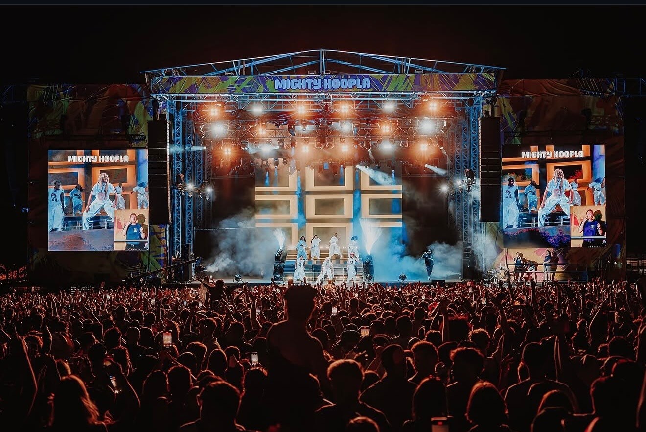 A packed crowd at Mighty Hoopla festival at night, arms raised, facing a brightly lit main stage. Performers dressed in white are visible on stage amid smoke and spotlights, with large LED screens on either side displaying the Mighty Hoopla branding and live footage of the performance.