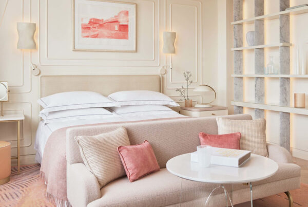 This is an image of a bright bedroom in the hotel One Aldwych. It has a beautiful double bed in its centre with a plush sofa at the end of the bed. The room is toned in neutral colours with muted pink highlights such as the cushions on the sofa. There are delicate light fixtures on the wall and a pink artwork on the wall above the bed.