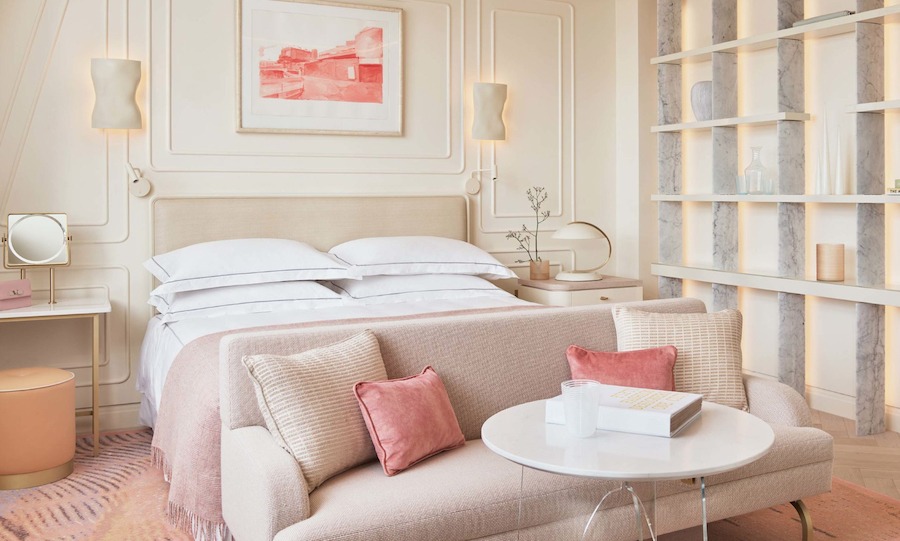 This is an image of a bright bedroom in the hotel One Aldwych. It has a beautiful double bed in its centre with a plush sofa at the end of the bed. The room is toned in neutral colours with muted pink highlights such as the cushions on the sofa. There are delicate light fixtures on the wall and a pink artwork on the wall above the bed.