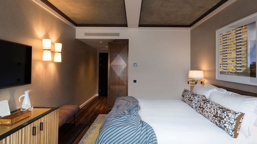 This is an image of a bright hotel bedroom. There is a large double bed in the centre of the room with a television hung on the opposite wall. There is soft warm lighting in the room and a neutral palette. The floor is wooden and the table opposite the bed is also made of wood. 