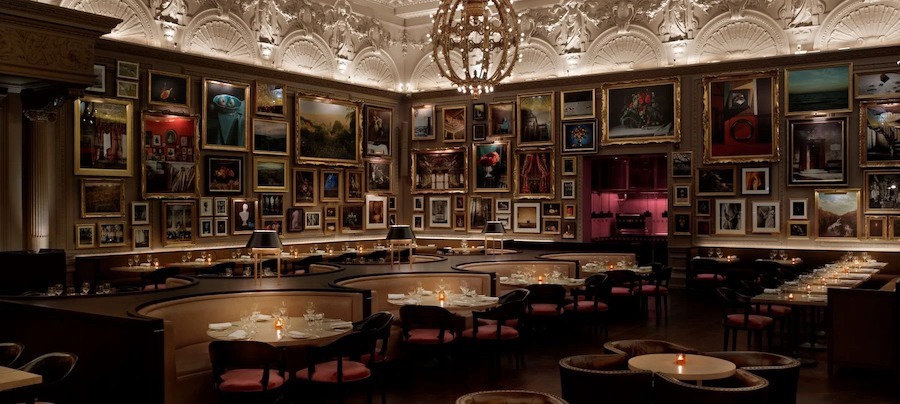 This is an image of a dimly lit restaurant. There are lots of semi circle booths around the room that are empty. On the walls of the restaurant hundreds of paintings and artworks are packed together giving the room a decadent and luxurious appearance. The ceiling and architectural features are ornate and traditional.