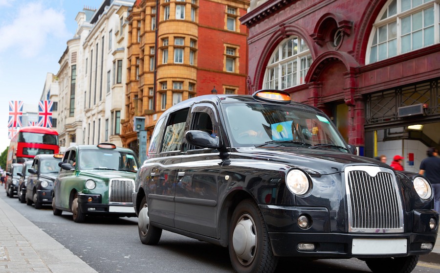This is an image of several London cabs driving down a street in London. The cars are clean and shiny and look well presented. There are UK flags hanging above a red bus in the background of the photograph. 
