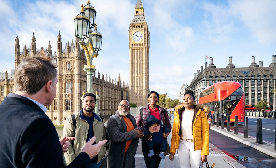 This is an image of a tour in London. A tour guide has his back to the camera and is giving a tour to a group of tourists who are smiling and looking happy. Big Ben is in the background and the sky is blue. 