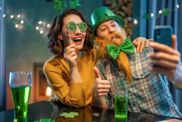 This is an image of a couple celebrating St Patrick's day. They are wearing funny green costumes such as a bow tie, hat, funny glasses and a headband. They are taking a picture together with a phone and have bright green drinks on the table in front of them.