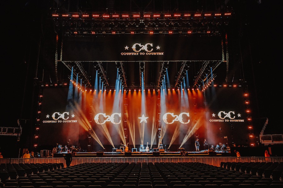 This i an image of a stage with dramatic spotlights shining on the stage. There is the lettering CC lit in various points on the stage and TV Screens either side of the stage.