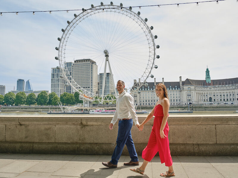 This is an image of a man and a woman walking hand in hand by a bridge. In the background, London Eye is seen in it's full height and width. The woman is wearing a bright red jumpsuit and the man is wearing a smart shirt and blue trousers. They are looking towards the camera and are smiling as they do their London photo session.