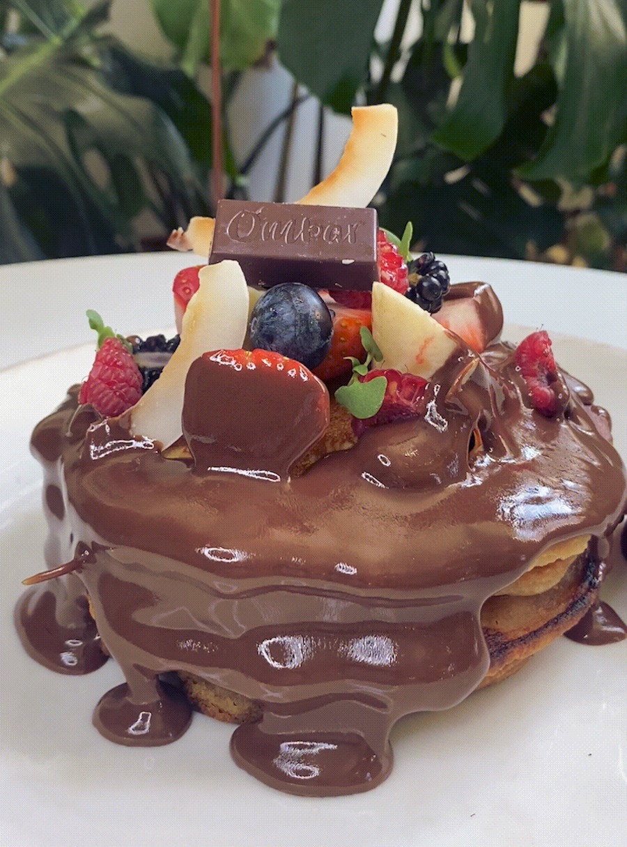 This is a closeup image of chocolate pancakes on a white plate covered with chocolate sauce, strawberries, blueberries and a little chocolate bar on top.