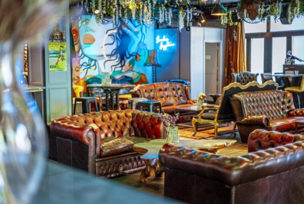 This is an image of a wheelchair accessible bar in London. There are various brown and red leather sofas around the room and funky cartoon art painted on the back wall. It has a cool and eclectic appearance to it. There is a neon blue sign at the back of the room and hanging plants on the ceiling.