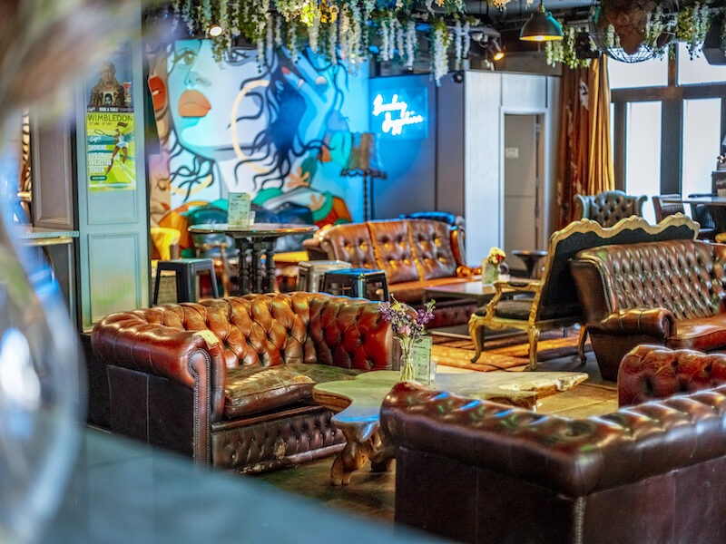 This is an image of a wheelchair accessible bar in London. There are various brown and red leather sofas around the room and funky cartoon art painted on the back wall. It has a cool and eclectic appearance to it. There is a neon blue sign at the back of the room and hanging plants on the ceiling.