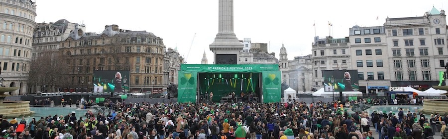 This is an image of a busy St Patrick's day gathering in London. There is a mass of people gathered together outside in front of a stage with a green banner.