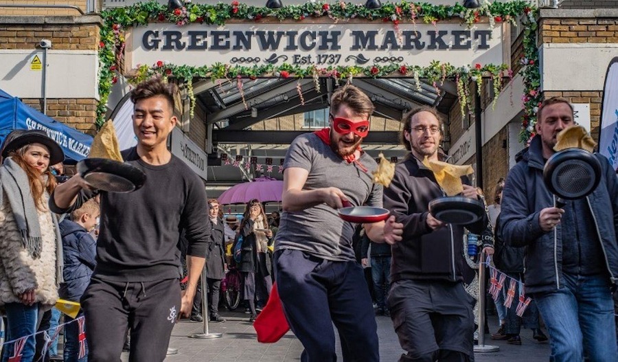 This is an image of a pancake race at Greenwich Market. There are several people holding pancake pans and flipping pancakes as they race together looking happy and smiling to celebrate Pancake day in London. 