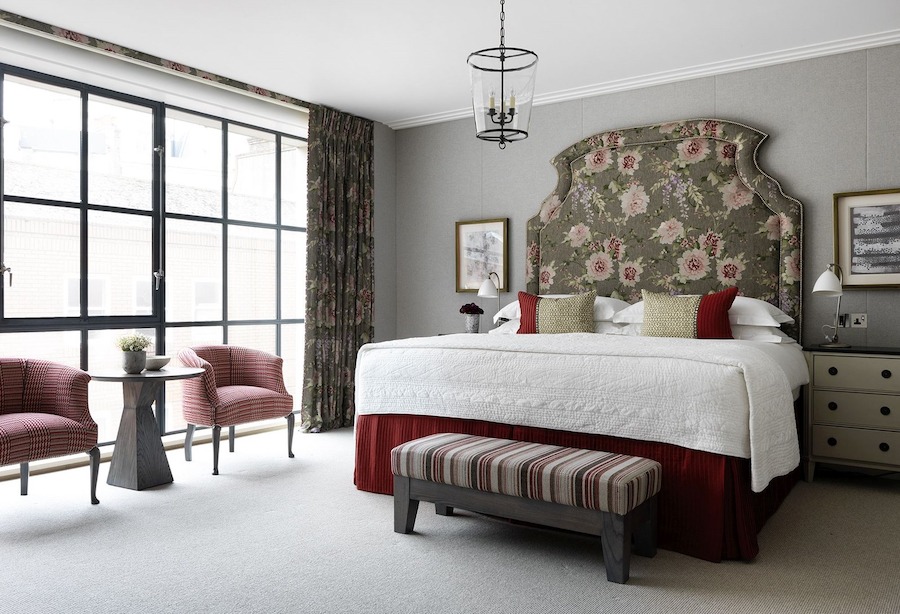 This is an image of a luxury hotel bedroom in London. In the middle of the room there is a neatly made double bed with white bedding with dark red touches. The floor is carpeted and clean, and the carpet, ceiling and walls are toned in neutral muted grey colours. There are two muted pink armchairs in front of the wall length windows on the left side of the image.
