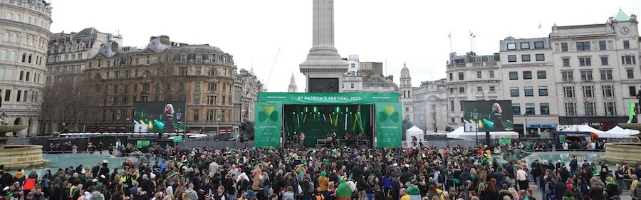 This is an image of the 2023 St Patrick's day festival in London. There are hundreds of people gathered together in celebration in central London. The sky is colourless and the buildings are grey. A bright green stand framed the central stage of the festivities.