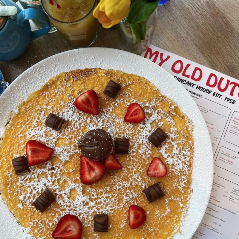 This is an image of a plate of pancakes with a menu underneath it on a wooden table with yellow roses in a glass vase next to it and a beige drink with a straw inside. On top of the pancakes there are little chocolate squares and slices of strawberries neatly laid out and powdered sugar sprinkled on top.