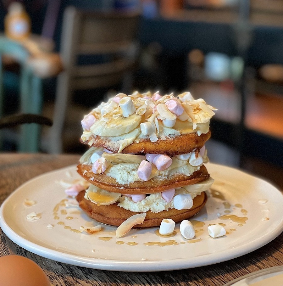 This is an image of three neatly stacked circular pancakes on a white plate. They are topped with marshmallows and cream that stick the pancakes together like a sandwich. 