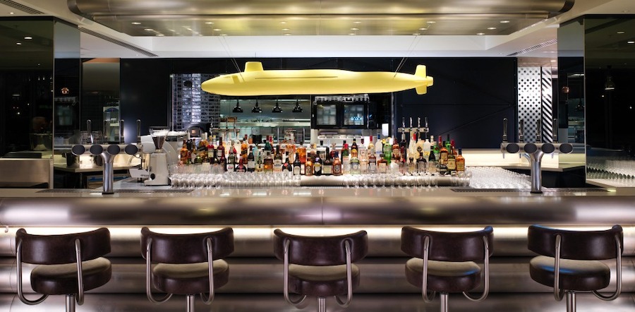 This is an image of a sleek, modern looking bar in a restaurant. There are small spotlights lighting the room giving it a dimly lit appearance. Bottles of alcohol line the back of the bar and are surrounded by a clean grey counter and modern seating.