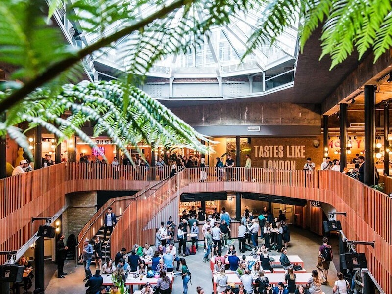 This is an image of Seven Dials Market. It is brightly lit and is full of people upstairs and downstairs dining at small tables or walking around and people watching. There is a bright skylight at the top of the ceiling and trees in the foreground.