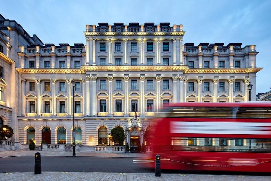 This is an image of a grand hotel. It is tall and wide and has a luxurious facade with beautiful archways and lighting features. There is a red bus driving in front of the hotel on the street opposite it and the sky is blue in the background.