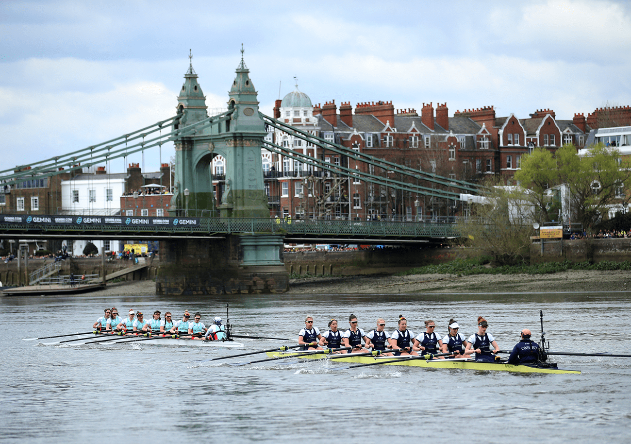 This is an image of a boat race across the River Thames in London. The sky is blue, the water is calm, and there are two race boats packed with racers inside.