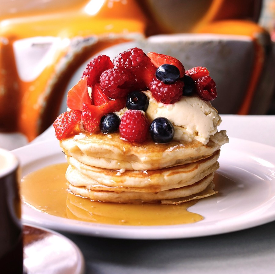 This is an image of neatly stacked pancakes on a plate. They are topped with vanilla ice cream, assorted berries and maple syrup.