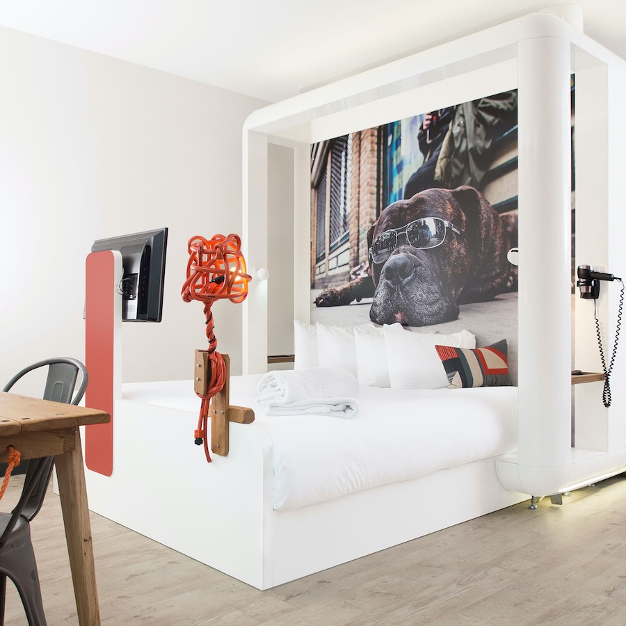 This is an image of a bright hotel bedroom with a large double bed in the middle of the room. There is a funny poster of a dog in sunglasses behind the head of the bed and a tv at the end of the bed. The walls and ceiling are white and the floor is wooden. 