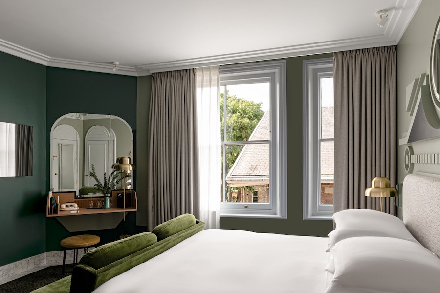 This is an image of a bright hotel bedroom with a neat double bed with white sheets and pillows. There is a big mirror on the opposite wall, two rectangular windows on the right hand side of the bed and muted green detailing to the neutral room. 
