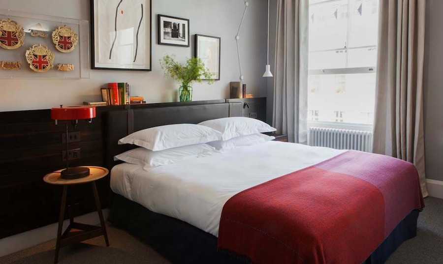 This is an image of a bright bedroom with a double bed in the middle of the room. The bed is neatly made with white sheets and a red duvet. On the wall behind the bed there are various sized artworks in contemporary frames. There is an open window beside the bed with stone coloured curtains. 