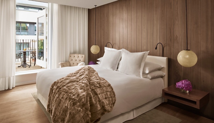 This is an image of a luxurious hotel bedroom. There is a neatly made double bed with white sheets and a fur throw in the middle of the room. The walls and floors are wooden and there are white curtains hanging either side of an open window on the right hand side of the bed.