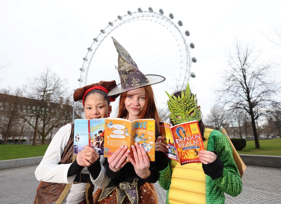 This is an image of three children reading books in front of the London Eye. They are wearing fancy dress costumes that reflect the books they are reading and look happy.