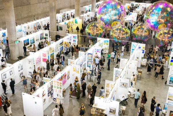 This is an image of a large room with high ceilings. Lots of people are looking at art on the white walls. There are many rows of white walls where artists work is hanging and there are big bubble sculptures hanging above.