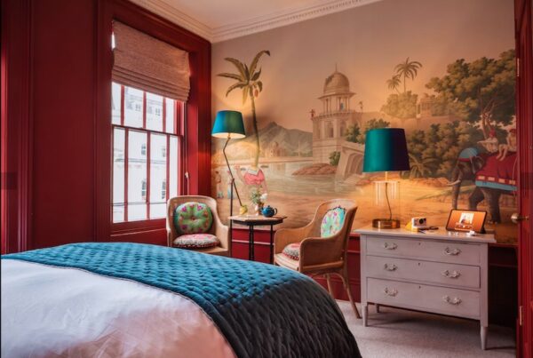 This is an image of a beautifully decorated hotel bedroom. There is a neatly made double bed in the middle of the room with white bedding and a dark turquoise cover. Opposite the bed is a wall featuring an artwork of a beach with camels and palm trees painted in a soft, figurative style. The floor of the room is carpeted and the walls are painted in a dark red colour.
