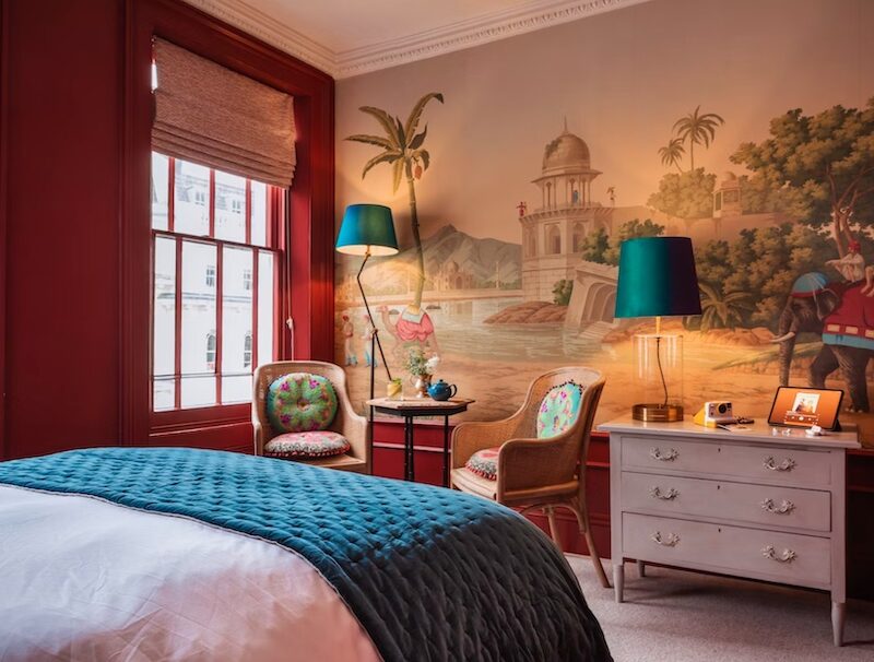This is an image of a beautifully decorated hotel bedroom. There is a neatly made double bed in the middle of the room with white bedding and a dark turquoise cover. Opposite the bed is a wall featuring an artwork of a beach with camels and palm trees painted in a soft, figurative style. The floor of the room is carpeted and the walls are painted in a dark red colour.