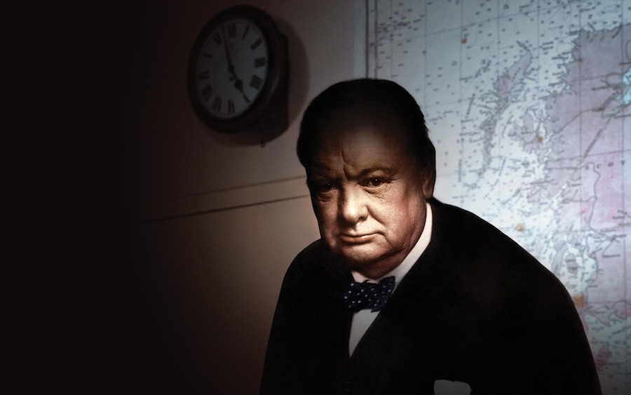 This is an image of Winston Churchill. He is dimly lit with a spotlight on his face. He is wearing a black suit and a bowtie and there is a map on the wall behind him.