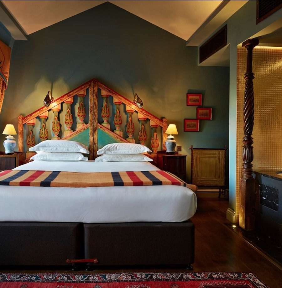 This is an image of a beautiful and whimsical hotel bedroom. There is a luxurious double bed with white sheets in the middle of the room. The walls are painted a muted green and the lighting is low and soft with lamps either side of the bed. The floor is a dark wood with a decadent red rug on top of it. 